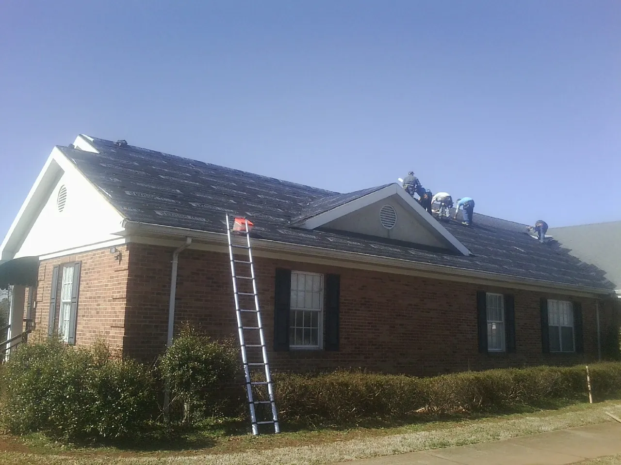 Crew tearing shingles off a brick ranch roof with a ladder set — Emergency Roof Repair work by our crew in Highland Lake