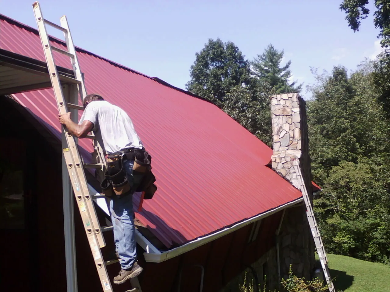 Roofer on a ladder beside a red metal roof and stone chimney — Soffit & Fascia Repair by Roofing Brothers in Highland Lake